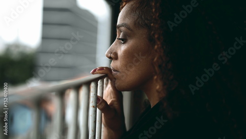 Profile close-up of a serious black woman leaning ln metal bar by home balcony gazing downwards with melancholic expression struggling but observing surroundings photo