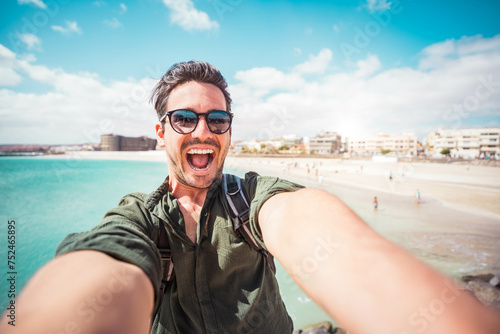 Handsome young man taking selfie with smart mobile phone device outside - Cheerful tourist enjoying summertime at the beach photo