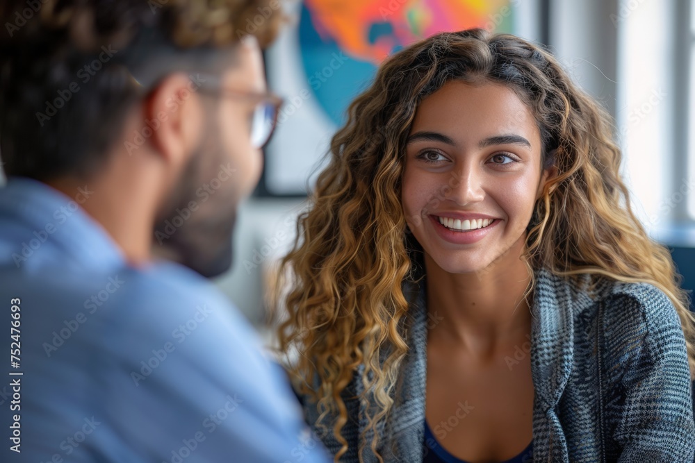 Obraz premium Joyful young woman engaging in conversation, radiant smile displaying confidence, person blurred in foreground. Exuberant female laughing in discussion, her curly hair and vibrant demeanor illustrate