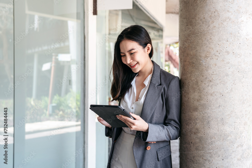 Asian businesswoman in suit, searching business data and reading text message on screen of digital tablet while standing Asian businesswoman in suit, searching business data and reading text message on screen of digital tablet while standing