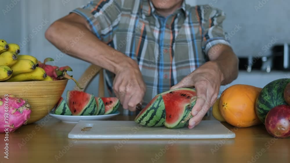 Young man surrounded by exotic tropical fruits cutting fresh red watermelon on a chopping board in the kitchen, slow motion. Healthy lifestyle and natural organic food concept Young man surrounded by exotic tropical fruits cutting fresh red watermelon on a chopping board in the kitchen, slow motion. Healthy lifestyle and natural organic food concept