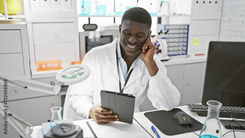 A smiling african man working in a laboratory with a tablet, magnifier, and computer equipment around. A smiling african man working in a laboratory with a tablet, magnifier, and computer equipment around.