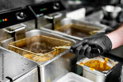 person in black gloves is frying food in professional kitchen using deep fryer. The stainless steel fryer contains bubbling oil, and adjacent containers suggest a busy commercial kitchen environment.