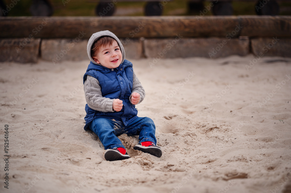 A young child dressed warmly in a vest and hat sits thoughtfully in the sand of a playground, capturing a moment of childhood tranquility and exploration A young child dressed warmly in a vest and hat sits thoughtfully in the sand of a playground, capturing a moment of childhood tranquility and exploration
