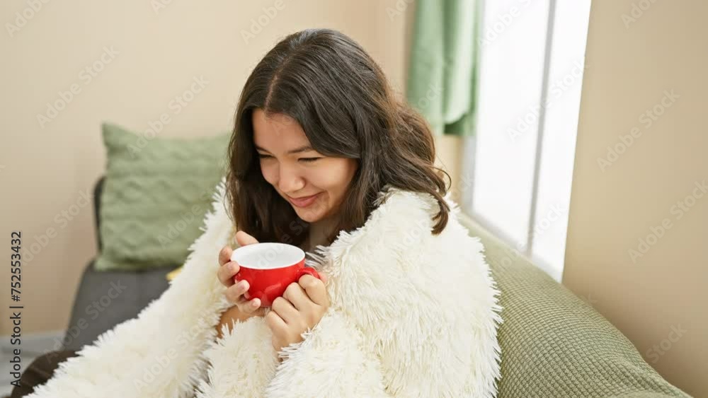 Hispanic woman in a cozy living room smiles while holding a red cup, depicting a relaxing moment at home. Hispanic woman in a cozy living room smiles while holding a red cup, depicting a relaxing moment at home.