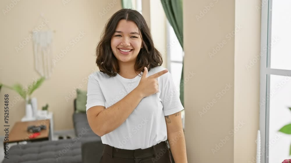 Cheerful young hispanic woman joyfully pointing up and to the side, bright smile on her face as she stands in the comfort of her beautiful home, invitingly gesturing towards something Cheerful young hispanic woman joyfully pointing up and to the side, bright smile on her face as she stands in the comfort of her beautiful home, invitingly gesturing towards something