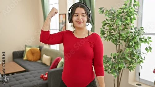 A young hispanic woman in a red top and headphones flexing her muscles with a smile indoors, depicting confidence and a healthy lifestyle in a living room setting. photo