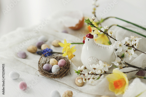 Easter still life. Colorful easter chocolate eggs in nest, spring flowers, chicken figurine composition on white wooden table. Space for text. Happy Easter! Seasons greetings