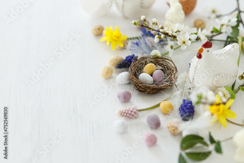 Happy Easter! Stylish easter chocolate eggs in nest, spring flowers, chicken figurine on white rustic wooden table. Easter modern simple banner, space for text. Seasons greetings