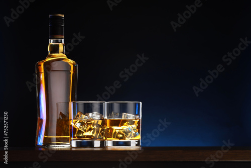 Whiskey with ice cubes in glasses and bottle on table against dark background, space for text © New Africa