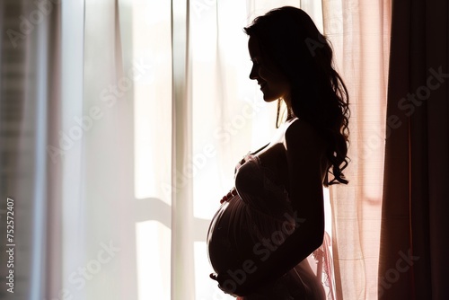 Happy pregnant woman in pink negligee standing by window looking at belly