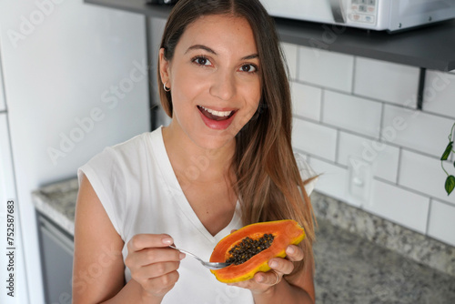 Portrait of beautiful woman holding half papaya fruit and spoon in the kitchen. Looks at camera.