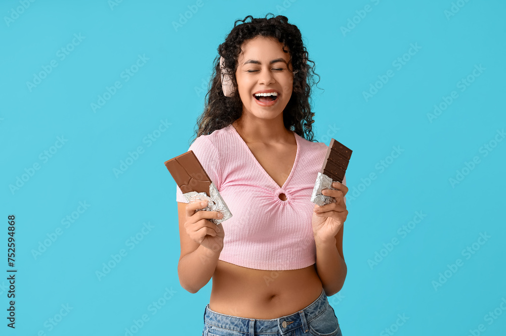 Beautiful young African-American woman in headphones with sweet chocolate bars on blue background - obrazy, fototapety, plakaty 