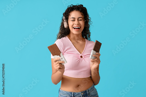Beautiful young African-American woman in headphones with sweet chocolate bars on blue background