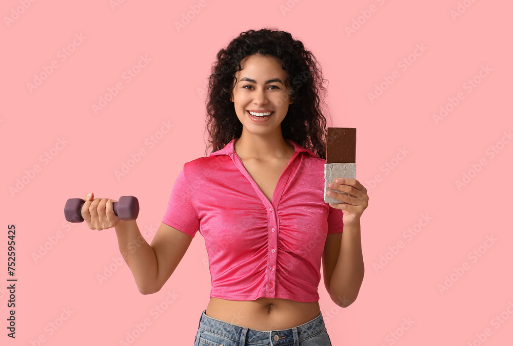 Happy young African-American woman with sweet chocolate bar and dumbbell on pink background - obrazy, fototapety, plakaty 
