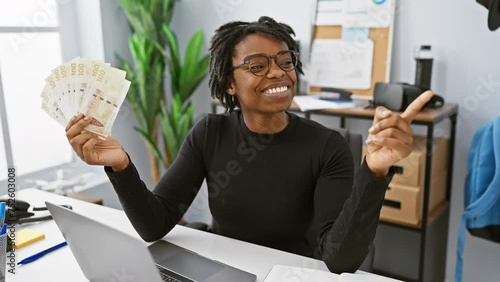 Joyous young black woman with dreadlocks in office, proudly brandishing danish krone banknotes. with a beaming smile and confident gesture, she points to one side. photo