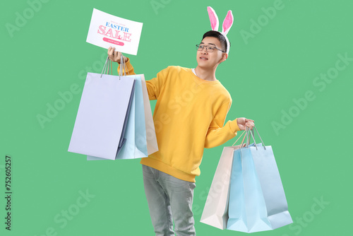 Young Asian man in bunny ears with shopping bags on green background. Easter Sale