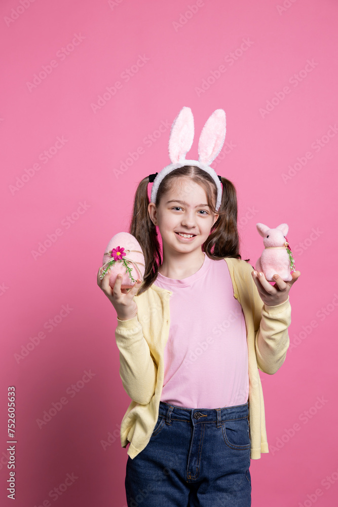 Joyful toddler presenting a pink egg and a stuffed rabbit in studio, wearing pigtails and cute bunny ears on camera. Smiling young kid showing her easter decorations and colorful toys. Joyful toddler presenting a pink egg and a stuffed rabbit in studio, wearing pigtails and cute bunny ears on camera. Smiling young kid showing her easter decorations and colorful toys.