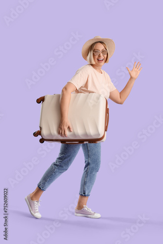 Young female tourist with suitcase waving hand on lilac background
