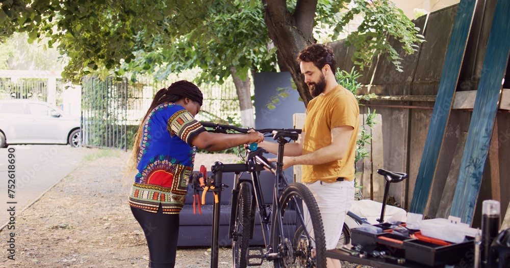 Obraz premium Sports-loving multiethnic couple fastening bike body for examination and maintenance. African american girlfriend assisting boyfriend by clutching modern bicycle to repair-stand in home yard.