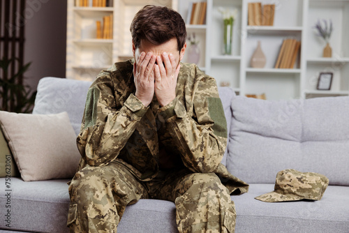Man in army clothing, home but facing depression. He is sitting on sofa, covering face with hands