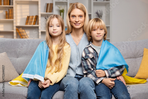 Beautiful mother embracing her children under Ukrainian flag, sitting together on living room sofa