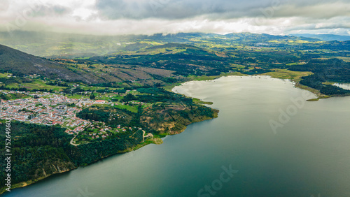 Fototapeta Naklejka Na Ścianę i Meble -  Aerial view of serene Guatavita Lake, lush greenery, quaint town, and mountains under a cloudy sky in Cundinamarca, Colombia
