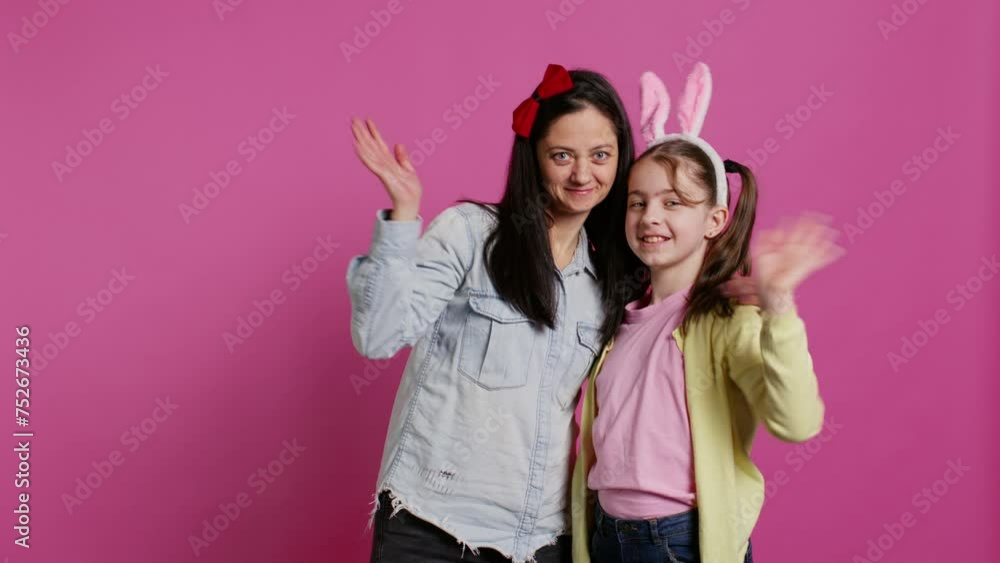 Lovely schoolgirl with bunny ears an her mom waving on camera, having fun and laughing against pink background. Cheerful mother and her daughter embracing and kissing each other. Camera B. Lovely schoolgirl with bunny ears an her mom waving on camera, having fun and laughing against pink background. Cheerful mother and her daughter embracing and kissing each other. Camera B.
