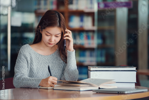 Unhappy young asian woman talking on mobile phone while reading book in library.