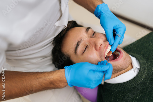 Portrait of young smiling man having teeth checked by dentist during appointment at dental clinic. Closeup of unrecognizable male dentist examining patient teeth with mouth mirror and dental excavator