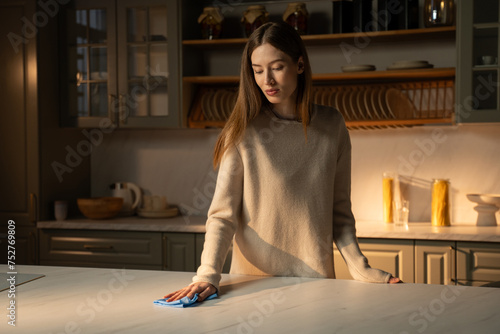 Canvastavla A focused young woman stands in a warmly lt kitchen, gently wiping the countertop with a blue cloth