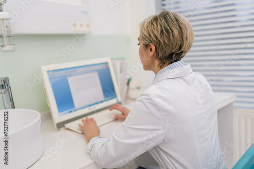 Rear view of unrecognizable middle-aged woman general practitioner in white coat using laptop computer writing notes at workplace. Female professional medic consulting patient distantly in online chat
