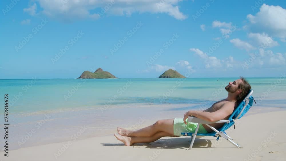 Side view of handsome bearded long-haired Caucasian guy in shorts relaxing in a folding chair on a beach with fascinating view of azure ocean and blue sky. High quality 4k footage Side view of handsome bearded long-haired Caucasian guy in shorts relaxing in a folding chair on a beach with fascinating view of azure ocean and blue sky. High quality 4k footage