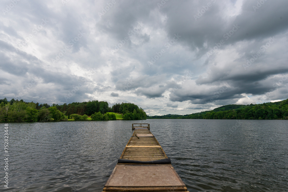Naklejka premium Dock on the lake. Wooden bridge in the forest in spring with blue lake on a cloudy day. Fishing lake with dock.