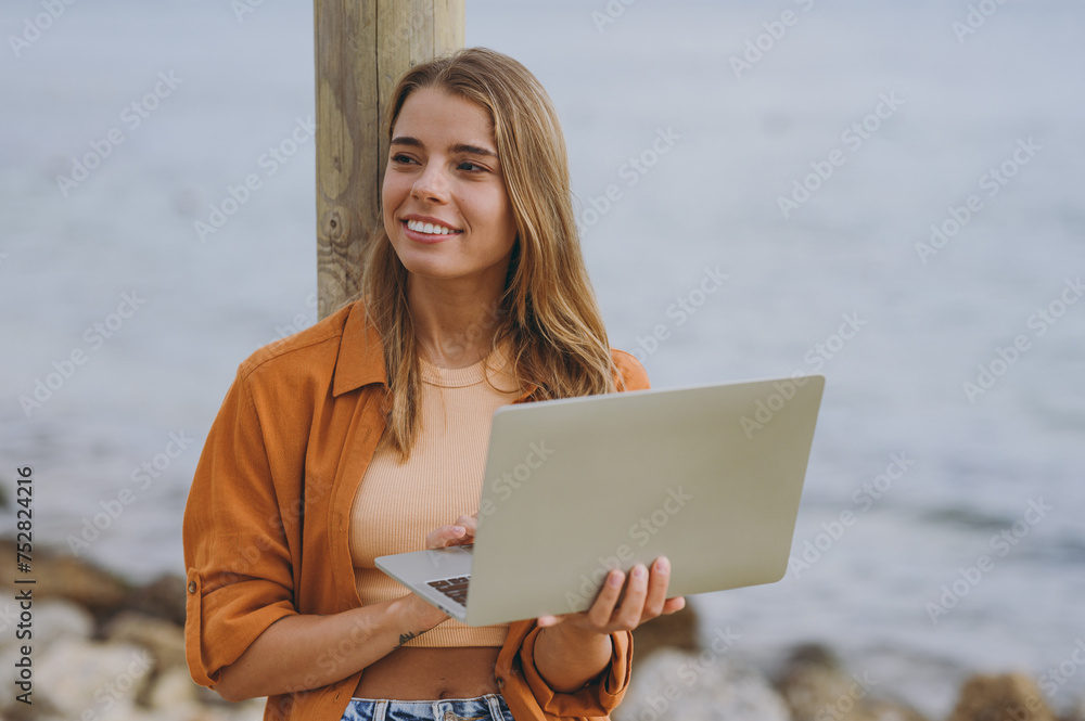 Young calm minded happy student IT woman she wear orange shirt casual clothes hold use work on laptop pc computer walk on sea ocean sand shore beach outdoor seaside in summer day. Lifestyle concept