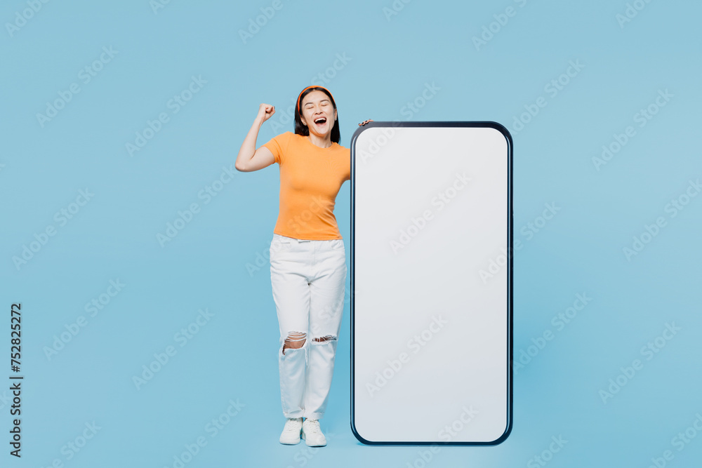 Φωτογραφία Young woman of Asian ethnicity in orange t-shirt casual clothes big huge blank screen mobile cell phone smartphone with workspace copy space mockup do winner gesture isolated on plain blue background