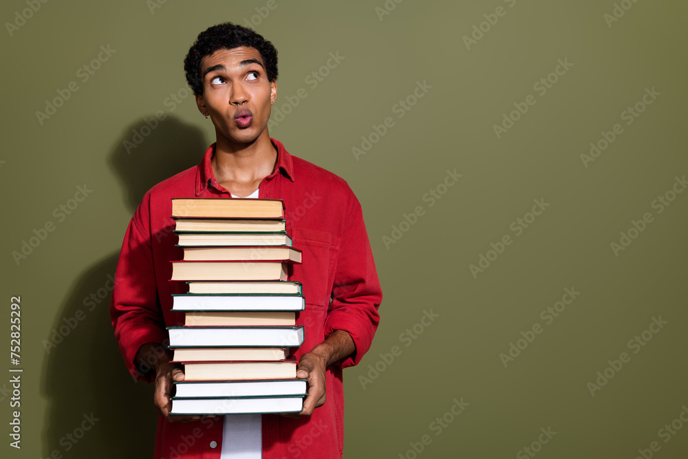 Photo of impressed uncertain man dressed red shirt carrying big heavy book pile looking empty space isolated green color background Photo of impressed uncertain man dressed red shirt carrying big heavy book pile looking empty space isolated green color background