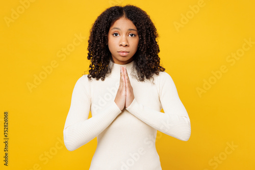 Little kid teen girl of African American ethnicity wear white casual clothes hold hands folded in prayer gesture, beg about something isolated on plain yellow background. Childhood lifestyle concept. photo