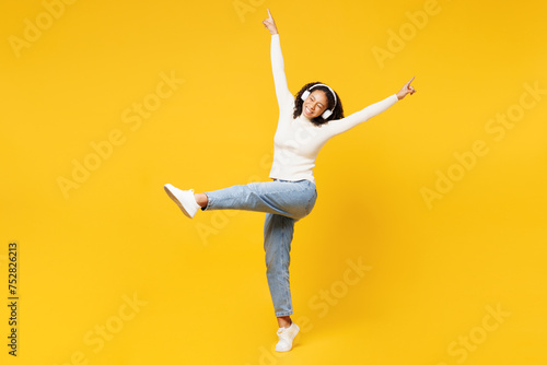 Full body cool little kid teen girl of African American ethnicity wear white casual clothes listen to music in headphones spread hands isolated on plain yellow background. Childhood lifestyle concept. photo