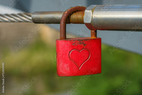 Love lock with carved heart hanging on the metal railing on the bridge. Green blurred background. Beautiful summer day.