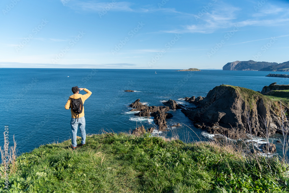 dark-haired man with yellow sweatshirt and backpack on his back looking out over the Basque coastline, holding his hand up to the sun.