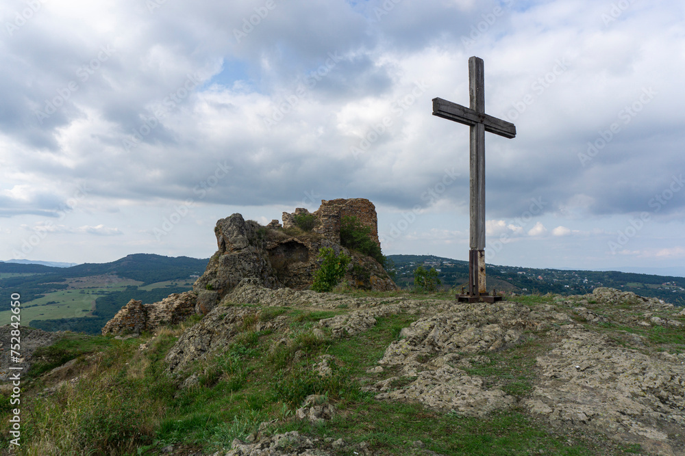 On the top of Kojori fortress. Ruins of the wall and tower made from rock and bricks. Tall wooden cross. Landscape of nearby mountains and villages. Clear blue sky and clouds. Georgia.