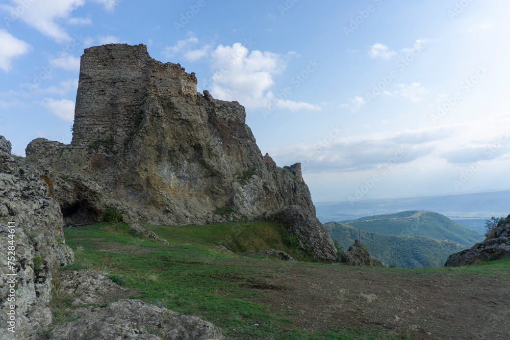 The ruins of a medieval fortress on top of a huge rock. Stone walls are a continuation and are combined with rocks.  Clear blue sky and clouds. Landscape of nearby mountains and villages. Kojori