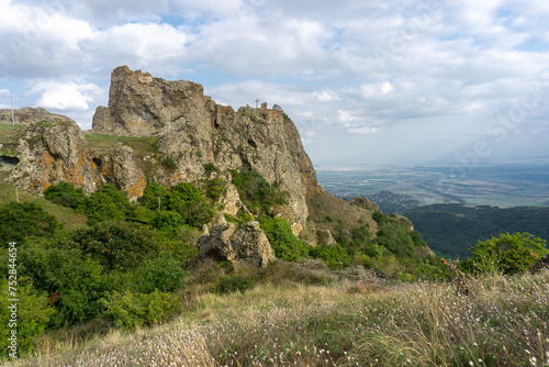 The ruins of a medieval fortress on top of a huge rock. Stone walls are a continuation and are combined with rocks. Clear blue sky and clouds. Landscape of nearby mountains and villages. Kojori