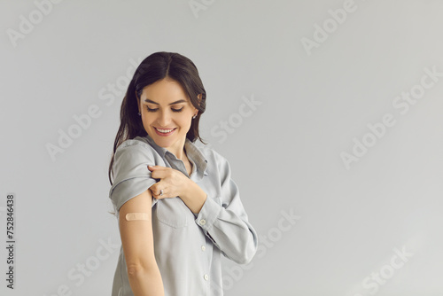 Happy smiling young lady showing arm after flu or Covid-19 vaccine injection. Woman promoting coronavirus vaccination and immunization campaign on light gray empty blank text studio background