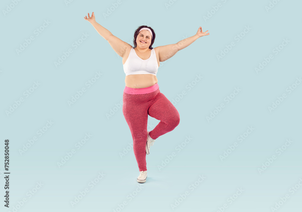 Fototapeta premium Happy cheerful joyful fat overweight woman in white sports bra and pink yoga pants isolated on light blue background enjoying fitness workout, doing exercise, standing on one leg. Weight loss concept