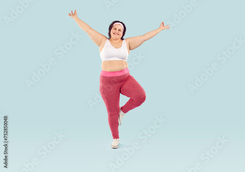 Happy cheerful joyful fat overweight woman in white sports bra and pink yoga pants isolated on light blue background enjoying fitness workout, doing exercise, standing on one leg. Weight loss concept © Studio Romantic