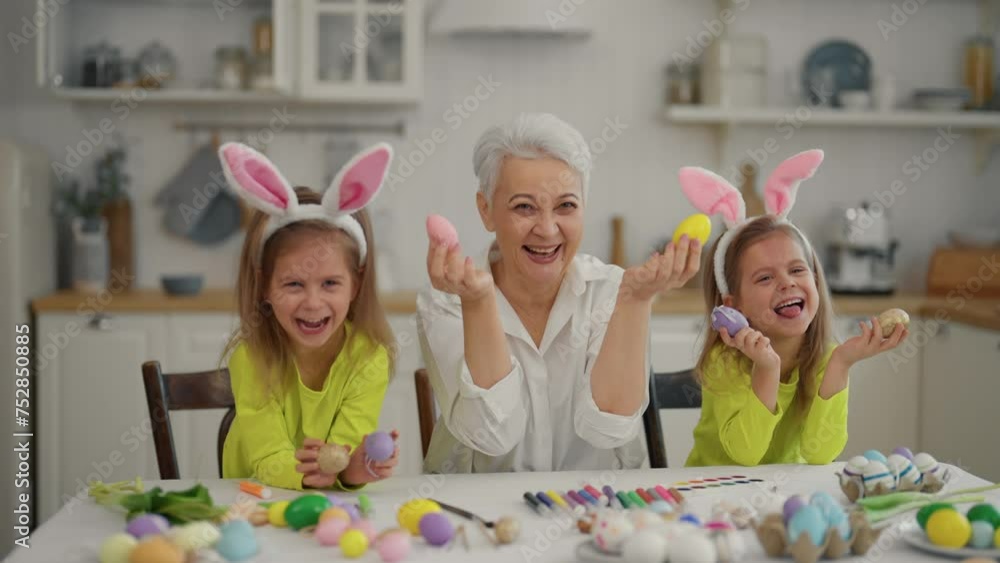 Easter grandmothers with granddaughters. Smiling grandmother with twins grandchildren wearing rabbit bunny ears decorating paint eggs together, show off painted eggs at kitchen. Family easter concept. Easter grandmothers with granddaughters. Smiling grandmother with twins grandchildren wearing rabbit bunny ears decorating paint eggs together, show off painted eggs at kitchen. Family easter concept.
