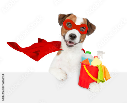 Happy jack russell terrier puppy wearing superhero costume holds bucket with washing fluids above empty white banner. Isolated on white background