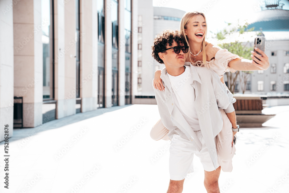 Young smiling beautiful woman and her handsome boyfriend in casual summer clothes. Happy cheerful family. Couple posing in street at sunny day. Female sits at his back. piggyback riding. Take selfie